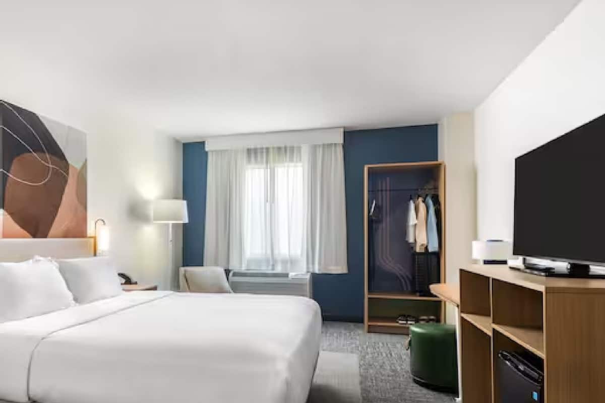 Una habitación de hotel con cama, mesitas de noche, televisor, armario y ventana con cortinas.