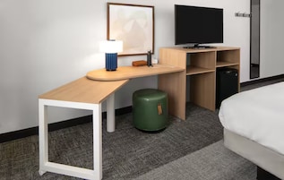 A hotel room with a bed, a desk with a lamp, a television, a green trash bin, and a wooden cabinet.