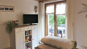 Una stanza con televisione su un mobile in legno, una libreria, una finestra con tende e una pianta.