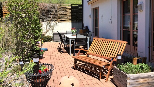 Un patio con una panchina di legno, un tavolo e sedie, circondato da piante in vaso e un giardino.