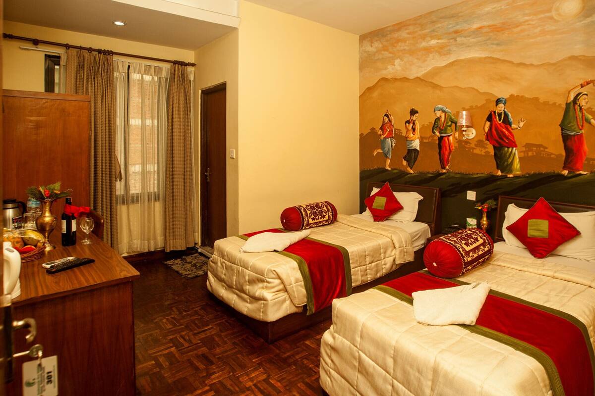 A hotel room with two beds, a wooden desk, a dining table with a vase and wine, and a mural on the wall.