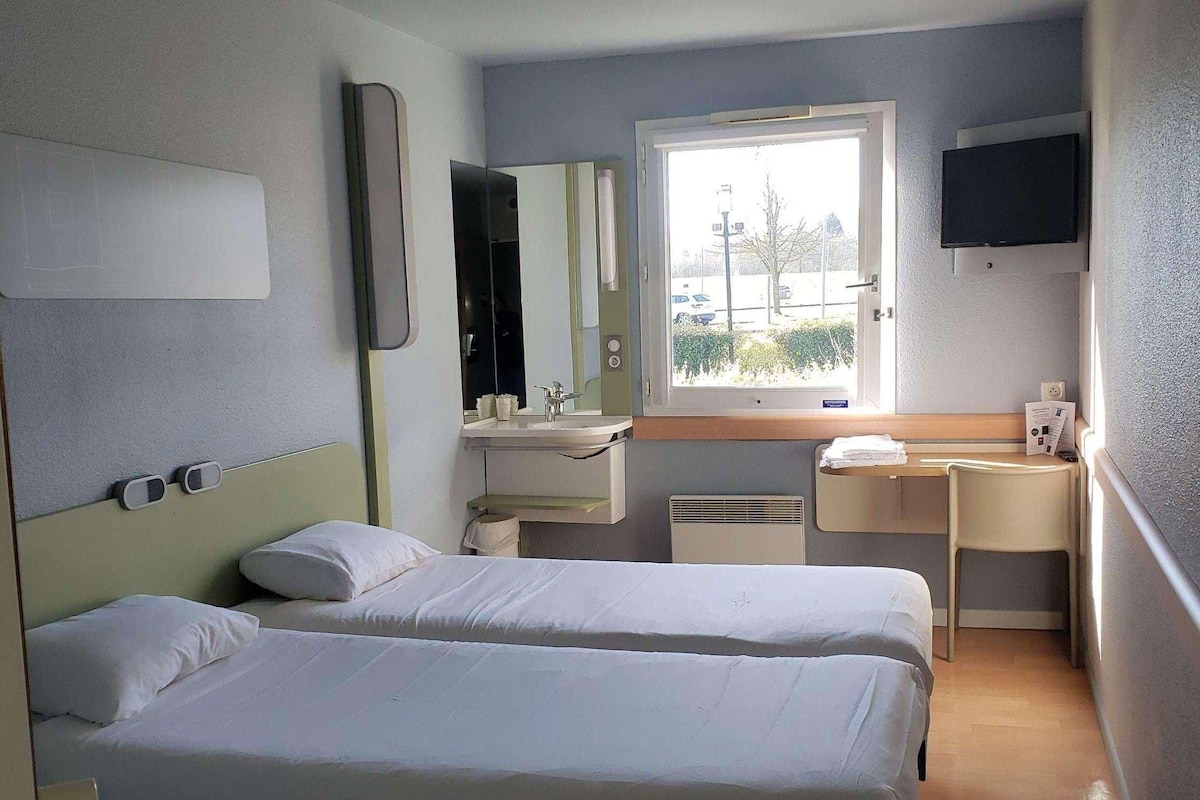 A hotel room with two beds, a desk, a TV, a window, and a bathroom.