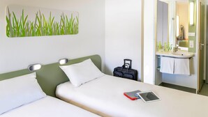 A hotel room with a bed, a bedside table with a lamp, a suitcase, and a bathroom with a sink and mirror. - Ibis Budget Dommartin les Cuiseaux (Dommartin-lès-Cuiseaux)