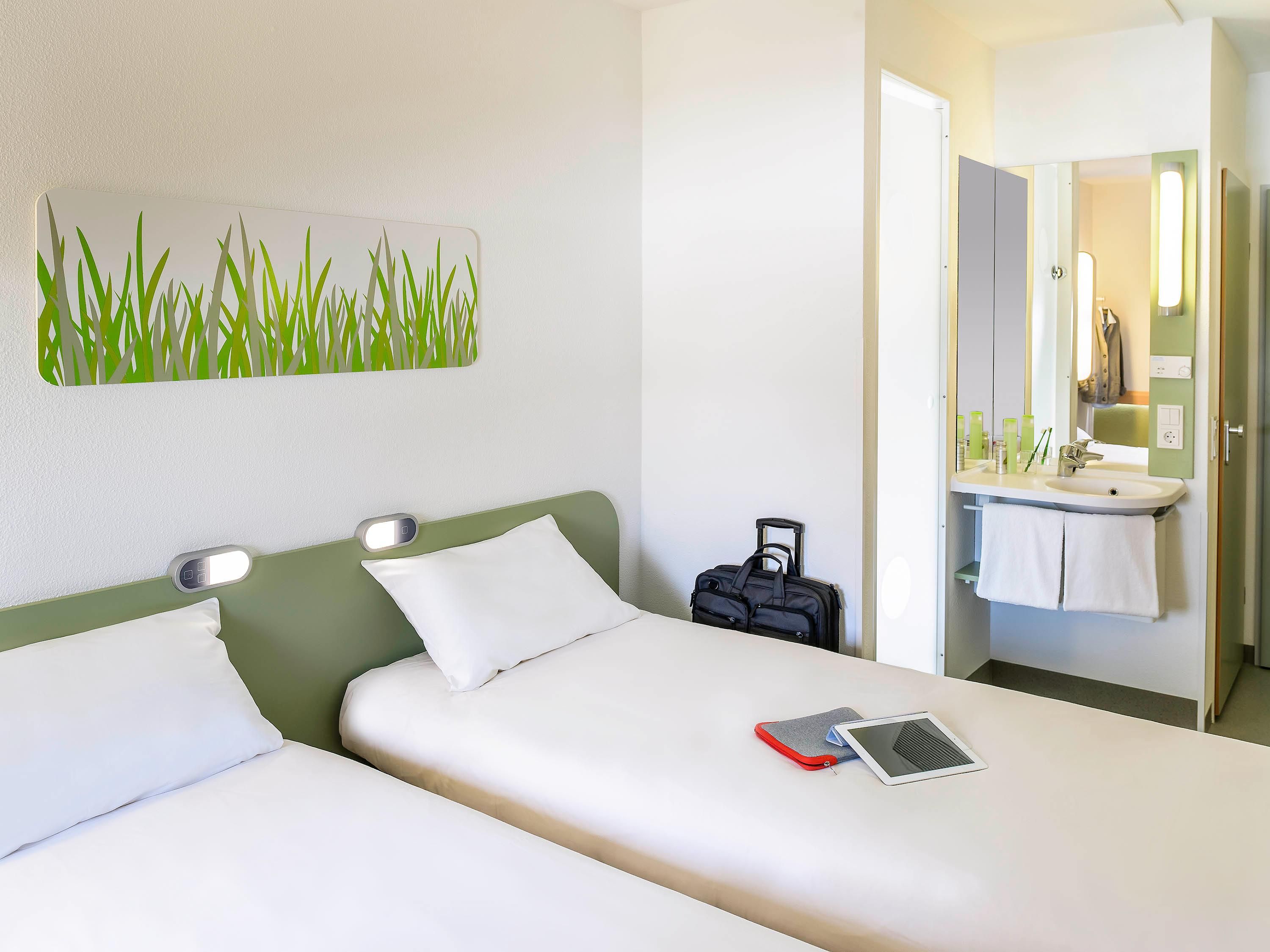 Una habitación de hotel con una cama, una mesita de noche con lámpara, una maleta y un baño con lavabo y espejo.