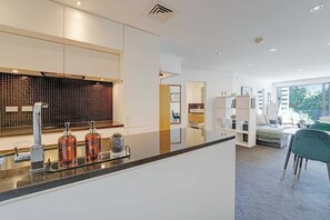 A modern kitchen with a large island, a dining area with a green chair, and a view of the outdoors. - Comfortable city fringe Apt Near Basque Park (Eden Terrace)
