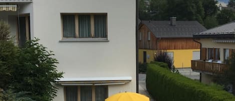 Ein zweigeschossiges Gebäude mit einer Terrasse. Dort steht ein Tisch mit Stühlen und einem gelben Sonnenschirm. Ein weißer Stuhl ist ebenfalls zu sehen.