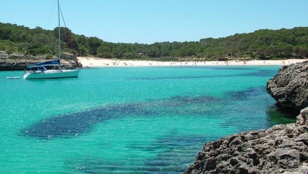 Escena costera con un velero cerca de una costa rocosa, una playa de arena y una colina boscosa al fondo.
