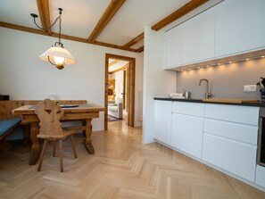 A wooden dining table with chairs, a kitchen with white cabinets, and a wooden ceiling with exposed beams. - FURTÜNADA, (Scuol) by Interhome (Scuol)