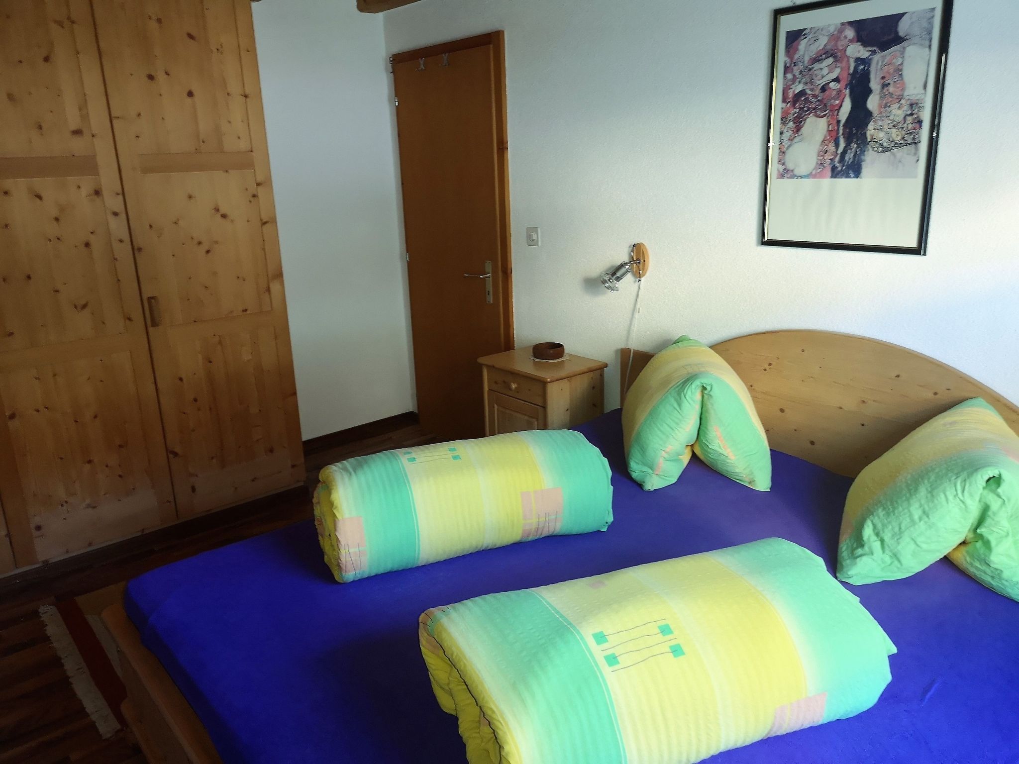 A room with a bed, a wooden wardrobe, a nightstand, and a wall-mounted lamp.
