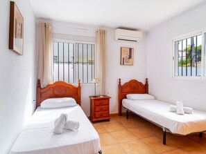 Ein Hotelzimmer mit zwei hölzernen Betten, einem Nachttisch, einer Klimaanlage und einem Fenster mit Blick auf Grünflächen.