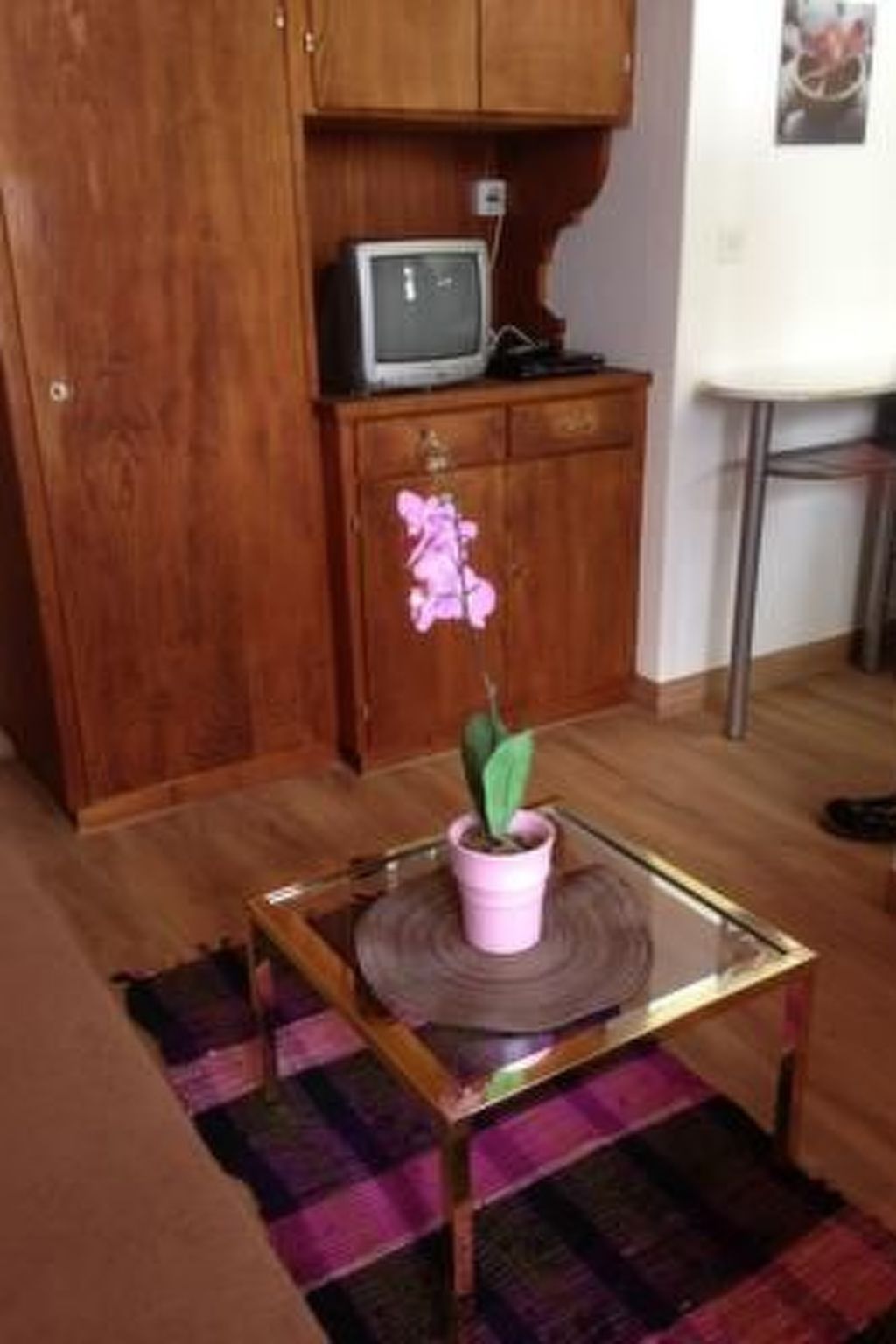 Ein Zimmer mit einem Holzkabinett, einem Fernseher darauf, einem kleinen Tisch mit einem Topfpflanzen und einem gemusterten Teppich.