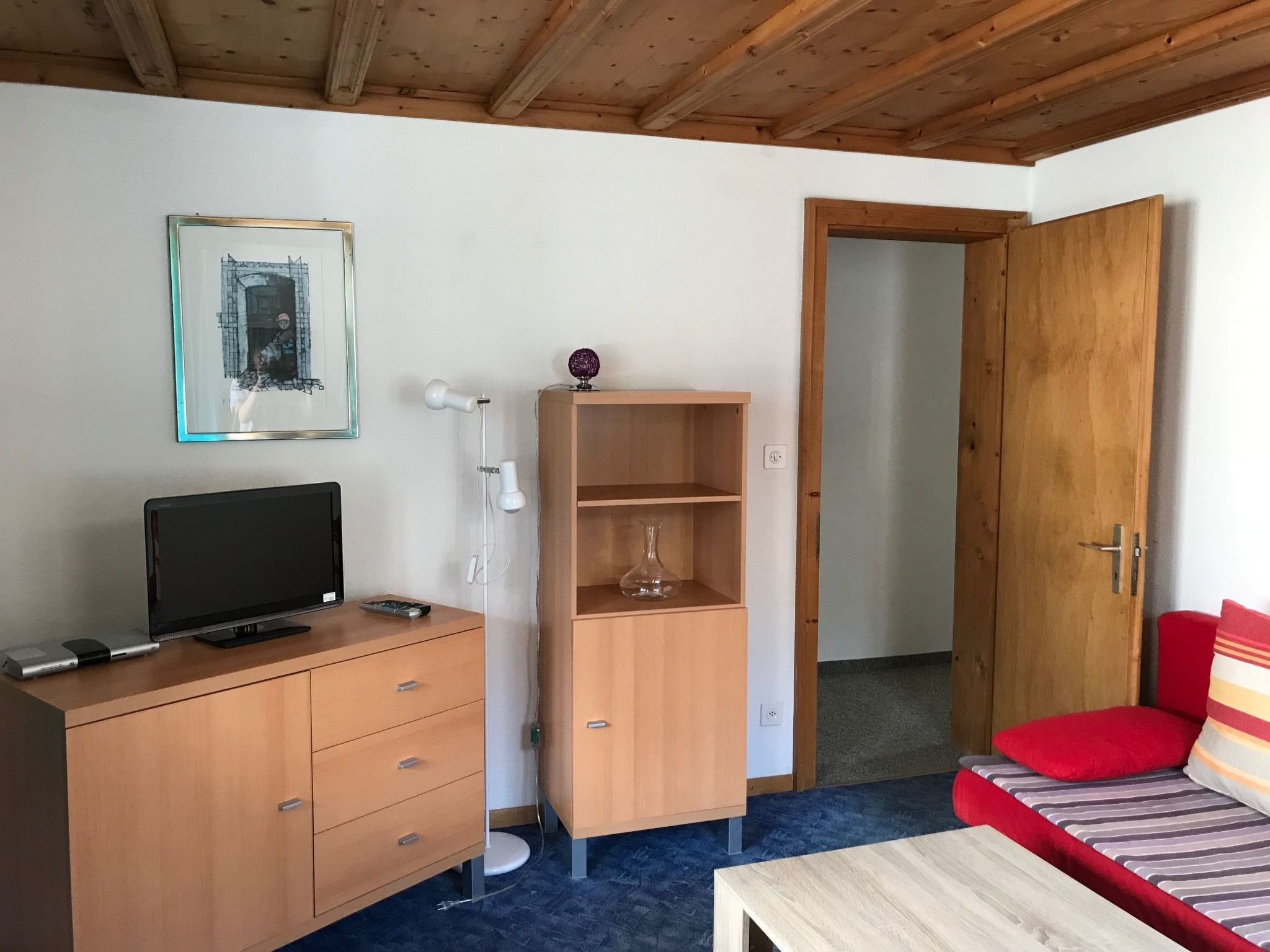 Een kamer met een televisie op een houten dressoir, een klein plankje met een vaas, een rode bank en een houten deur.