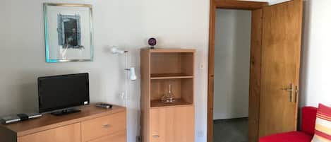 A room with a television on a wooden dresser, a small shelf with a vase, a red sofa, and a wooden door.