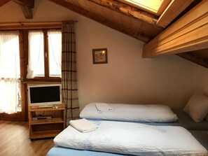 A room with a television on a wooden stand, two beds with white bedding, a window with curtains, and a picture on the wall. - Ferienwohnungen Chasa Valär, (Scuol) by Interhome (Scuol)
