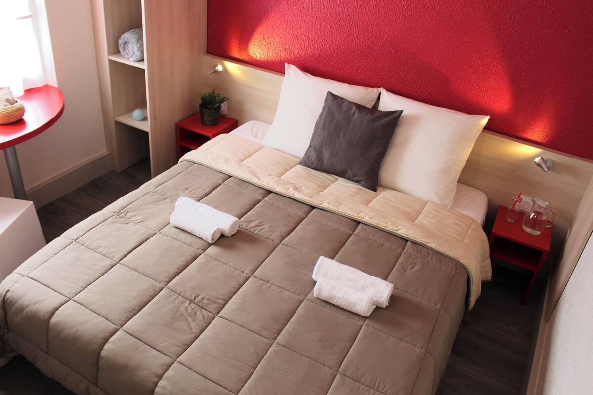 Une chambre d’hôtel avec un lit, une table de chevet, un mur rouge et une petite table sur laquelle est posée une plante.