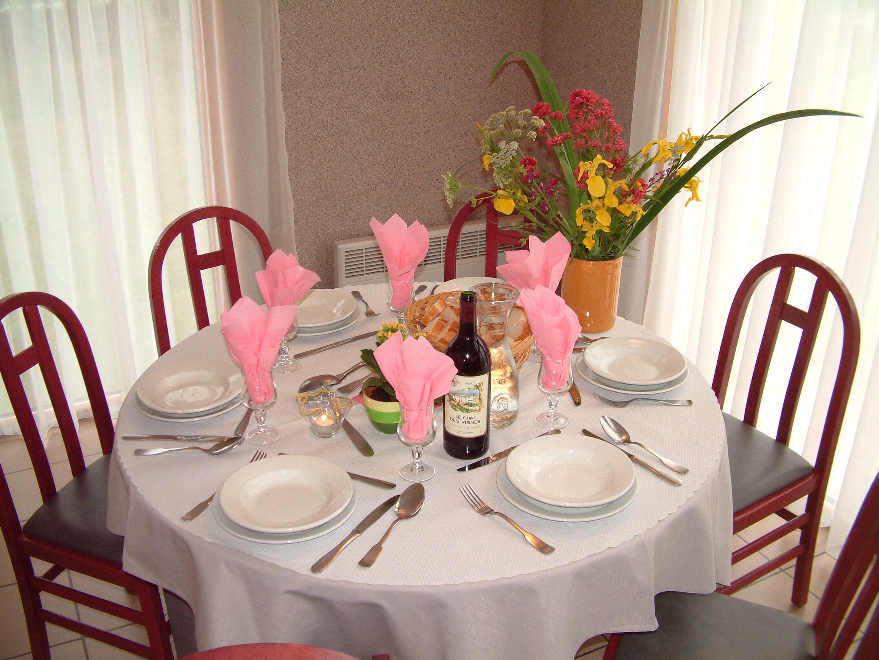 A round dining table set for four with white tablecloth, pink napkins, and a vase of flowers.