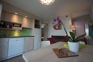 A modern kitchen with a dishwasher, microwave, and green-tiled backsplash. There's a dining area with a table, chairs, and a potted plant. - Vvf Villages Portbail (Port-Bail-sur-Mer)