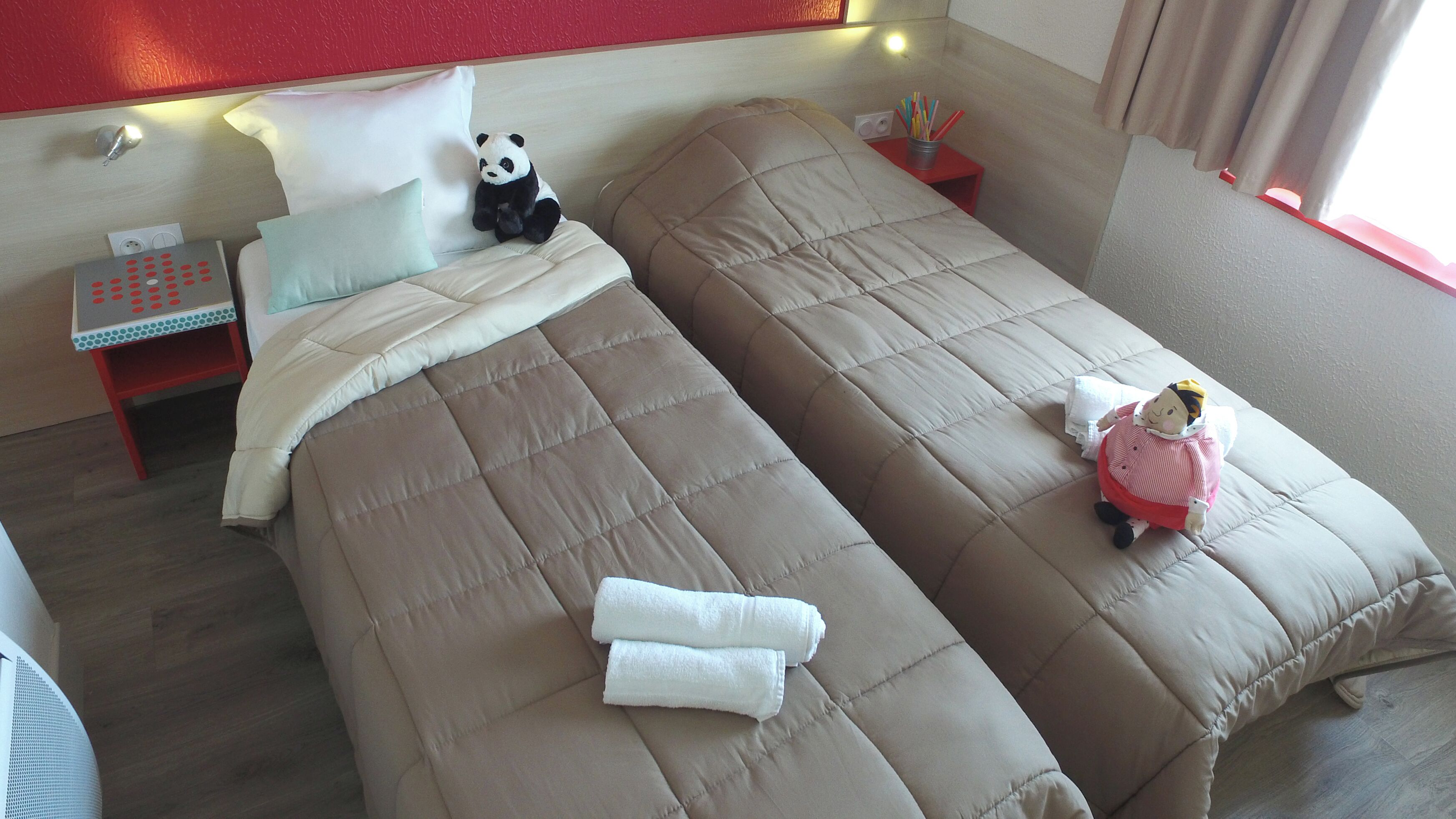 A hotel room with two beds, a nightstand, a red chair, a panda plush toy, and a doll.