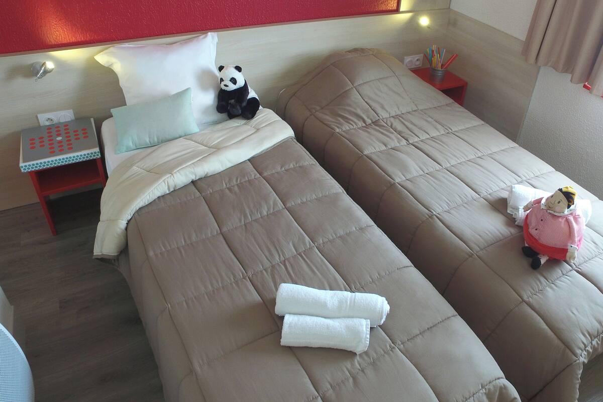 Une chambre d’hôtel avec deux lits, une table de chevet, un fauteuil rouge, un ours en peluche à l’effigie d’un panda et une poupée.