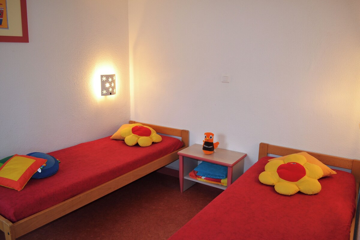Une chambre avec deux lits simples, chacun recouvert d’une courtepointe rouge et agrémenté d’un oreiller en forme de fleur. Une table de chevet avec une lampe se trouve entre les deux lits.