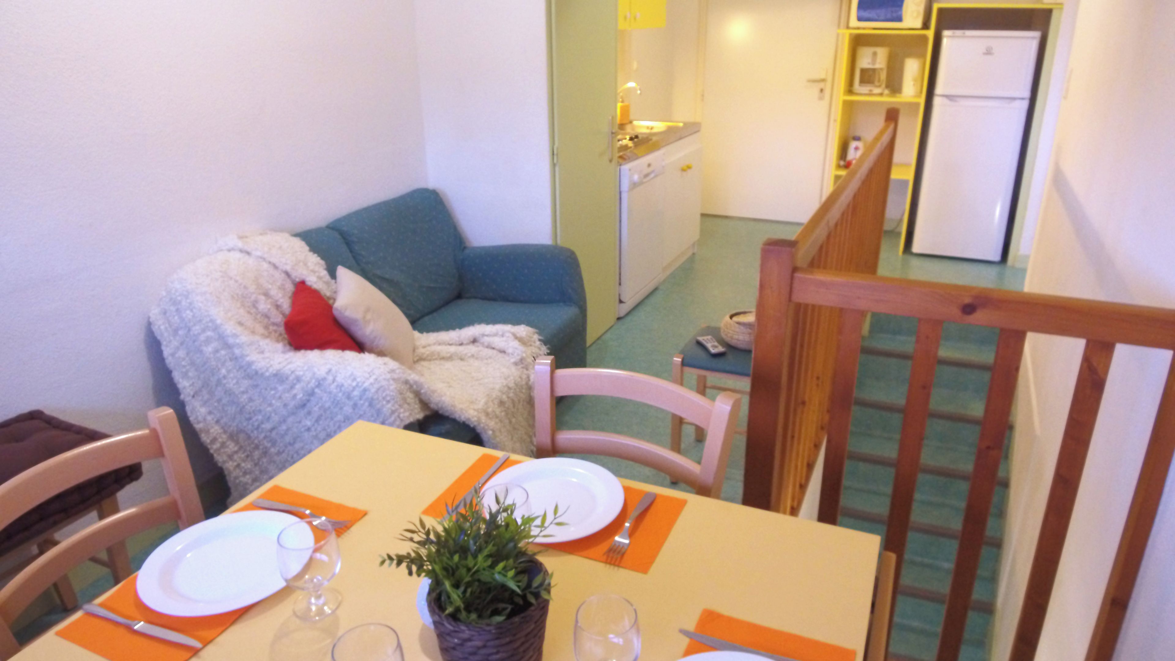 Uma área de refeições com uma mesa posta para dois, um sofá com manta e uma cozinha ao fundo.