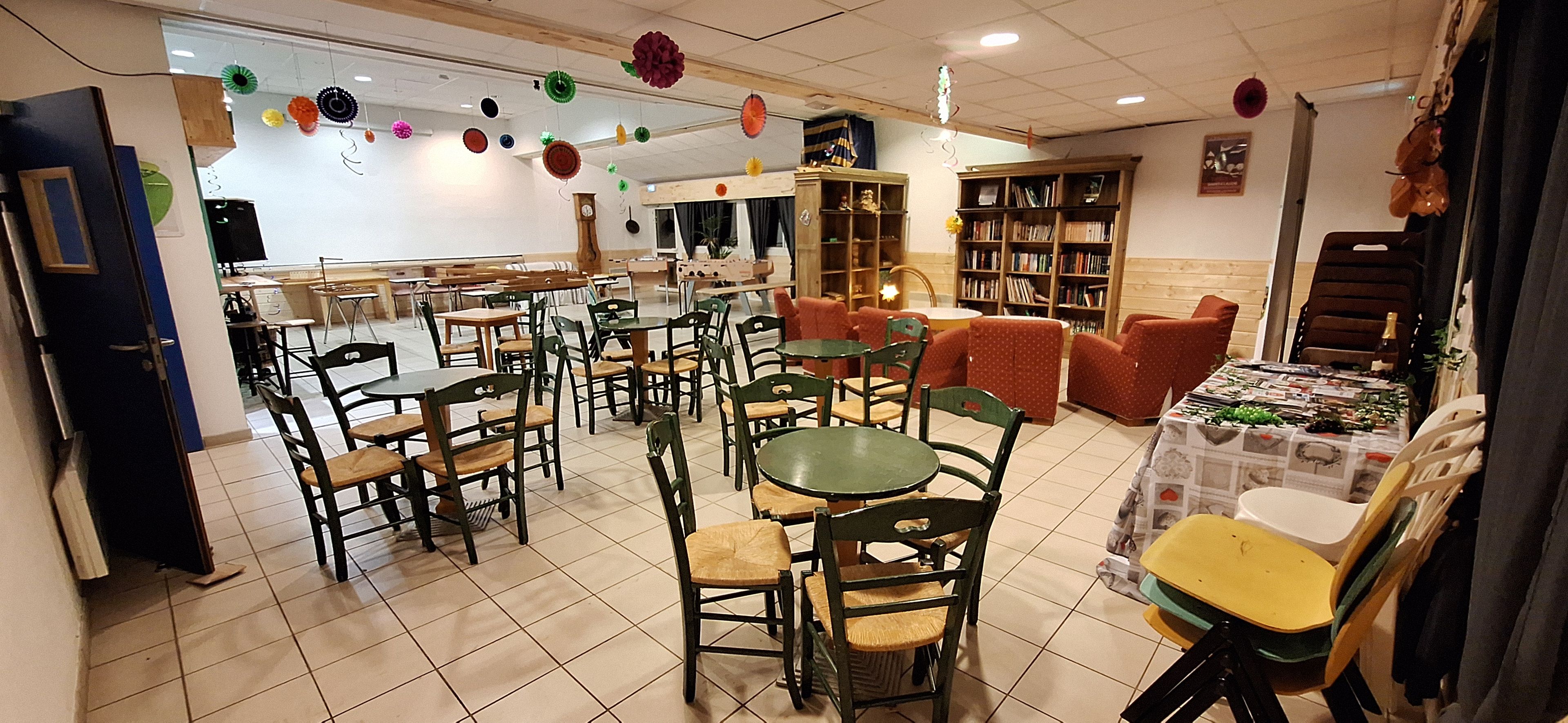 A dining area with multiple tables and chairs, a kitchen in the background, and a bookshelf filled with books.