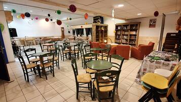 A dining area with multiple tables and chairs, a kitchen in the background, and a bookshelf filled with books.