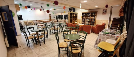 Un comedor con varias mesas y sillas, una cocina al fondo y una estantería llena de libros.