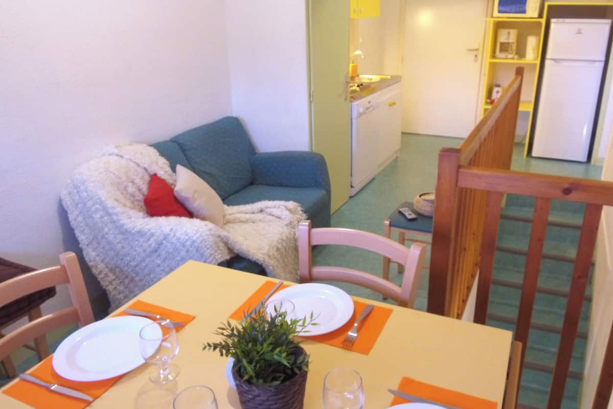 Ein Essbereich mit Tisch für zwei Personen, einer Couch mit Decke und einer Küche im Hintergrund.