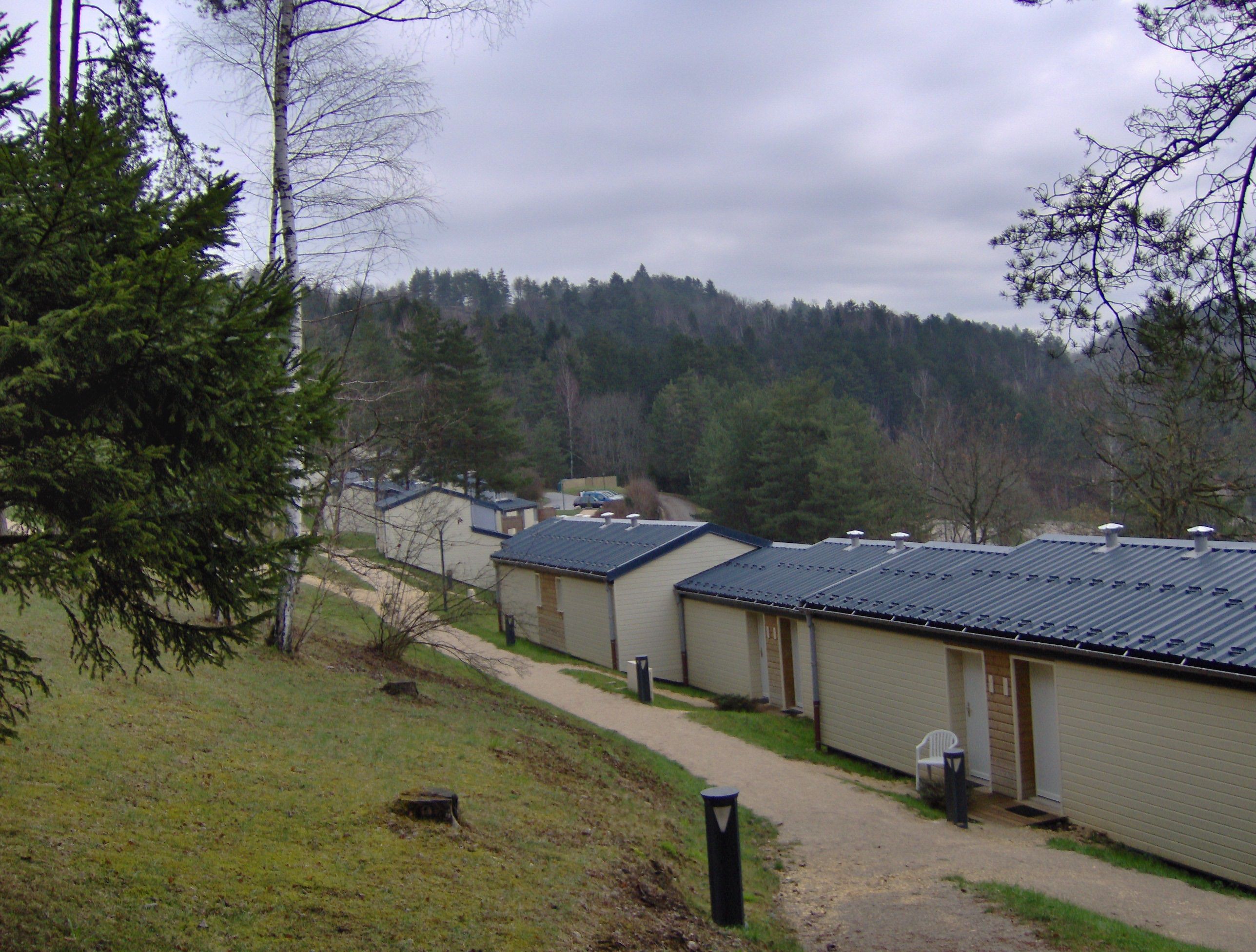 A row of single-story cabins with a dirt path leading to them, surrounded by trees and a forested hill in the background.