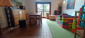 Una habitación con una mesa de comedor de madera, sillas, un pequeño espacio de juegos con bloques de colores y un cubo de basura.
