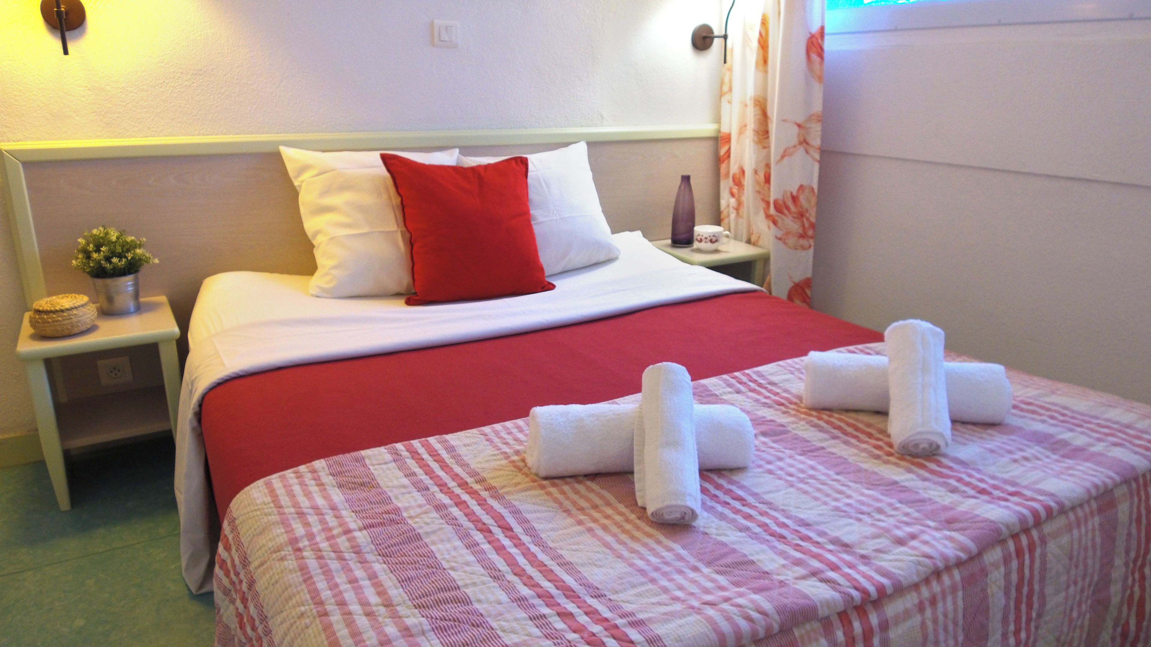 Una habitación de hotel con cama, mesita de noche, una pequeña planta en maceta y una ventana con cortinas.