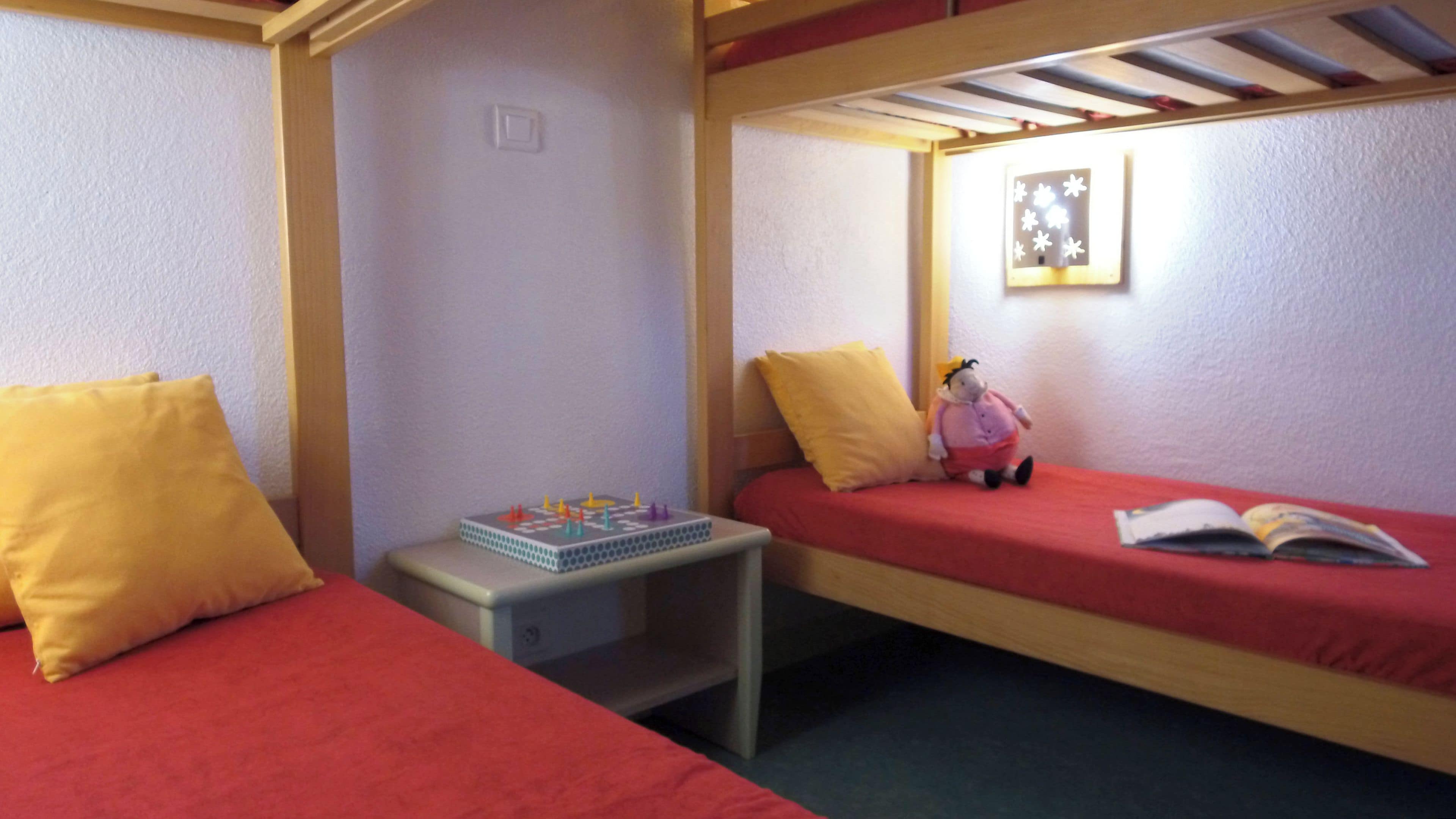Une chambre avec deux lits simples, une petite table avec un jeu de société, un jouet en peluche et un luminaire fixé au mur.