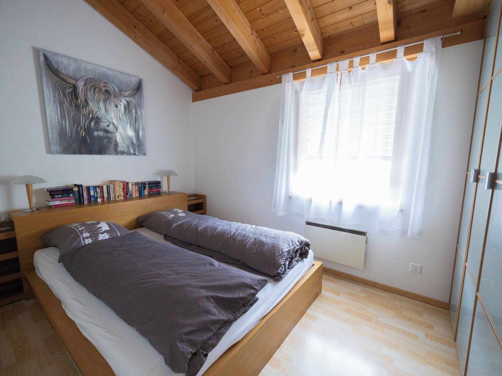 Une chambre à coucher avec un lit en bois, une table de chevet, une lampe, une fenêtre avec des rideaux et un tableau accroché au mur.