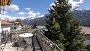 Ein Balkon mit Tisch und Stühlen, der auf eine Berglandschaft mit einem großen Nadelbaum und einem Gebäude mit rotem Dach blickt.