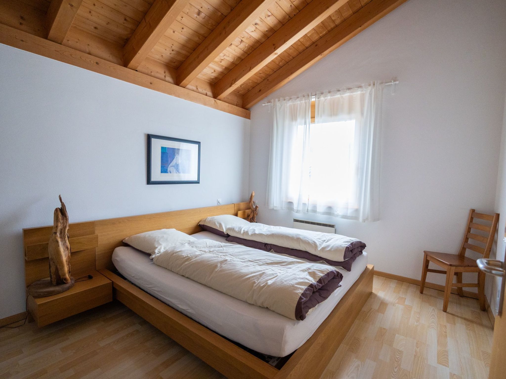 Ein Schlafzimmer mit Holzboden, einem Bett mit weißer Bettwäsche, einem Nachttisch aus Holz, einem Stuhl und einem Fenster mit Vorhängen.