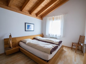 A bedroom with a wooden ceiling, a bed with white bedding, a wooden nightstand, a chair, and a window with curtains. - Senda 517c, (Scuol) by Interhome (Scuol)