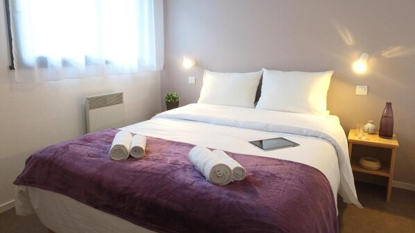 Ein ordentlich bezogenes Bett mit weißen Bettwäsche, einer violetten Tagesdecke und aufgerollten Handtüchern. Ein Nachttisch mit einer Vase und einer Lampe steht daneben.
