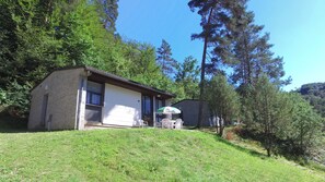 Een klein huis met een terras, omringd door bomen en een grasheuvel.