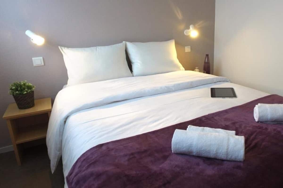 A neatly made bed with white linens, a purple blanket, and two rolled towels. There is a small bedside table with a plant and a bottle.