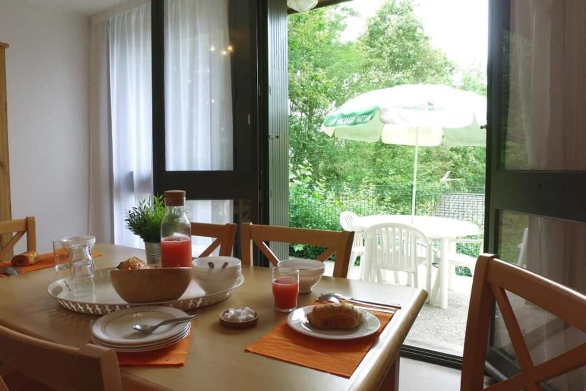 Une table à manger avec des aliments pour le petit-déjeuner, des chaises, une fenêtre donnant sur un paysage verdoyant et une porte.