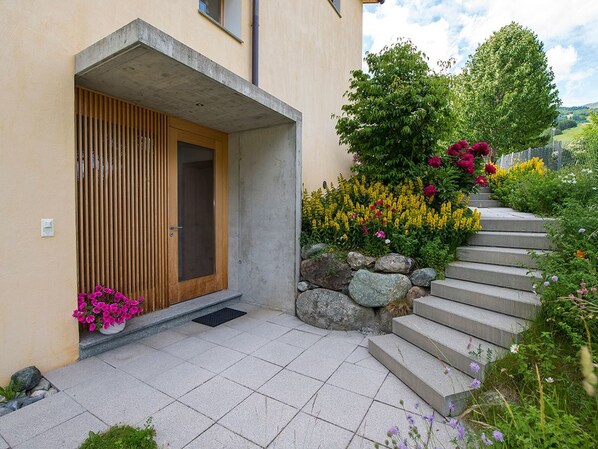 Un bâtiment moderne doté d’une porte en bois, d’un auvent en béton et d’un escalier en pierre menant à un jardin où poussent diverses fleurs et plantes.