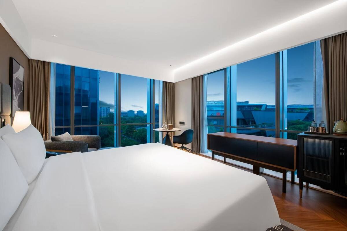 Une chambre d’hôtel moderne dotée d’un grand lit, d’un bureau, d’une chaise et offrant une vue sur le paysage urbain grâce à de grandes fenêtres.