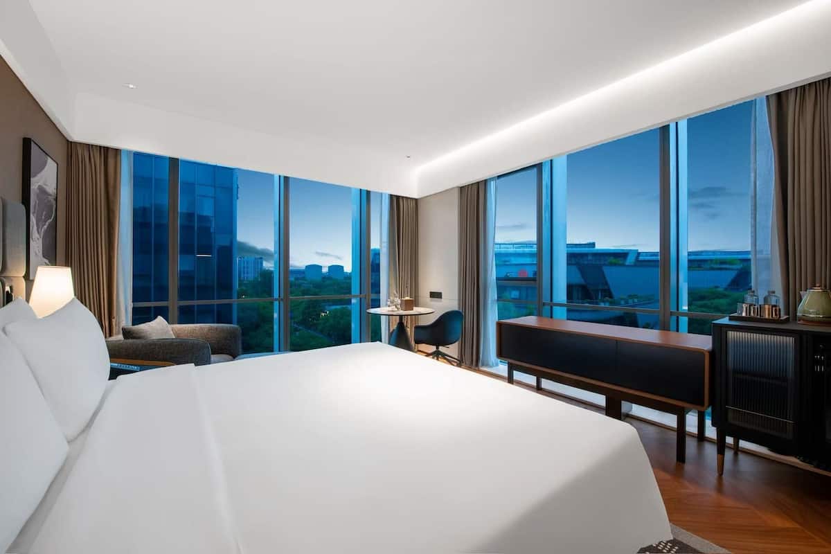A modern hotel room with a large bed, a desk, a chair, and a view of the cityscape through large windows.