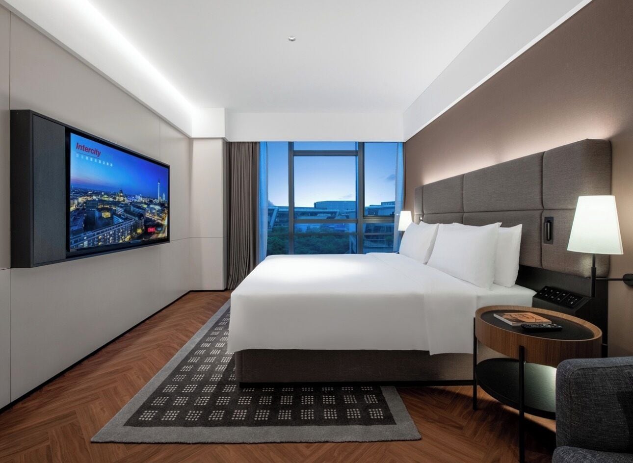Une chambre d’hôtel moderne avec un grand lit, un téléviseur fixé au mur, une table de chevet et une vue sur la ville.