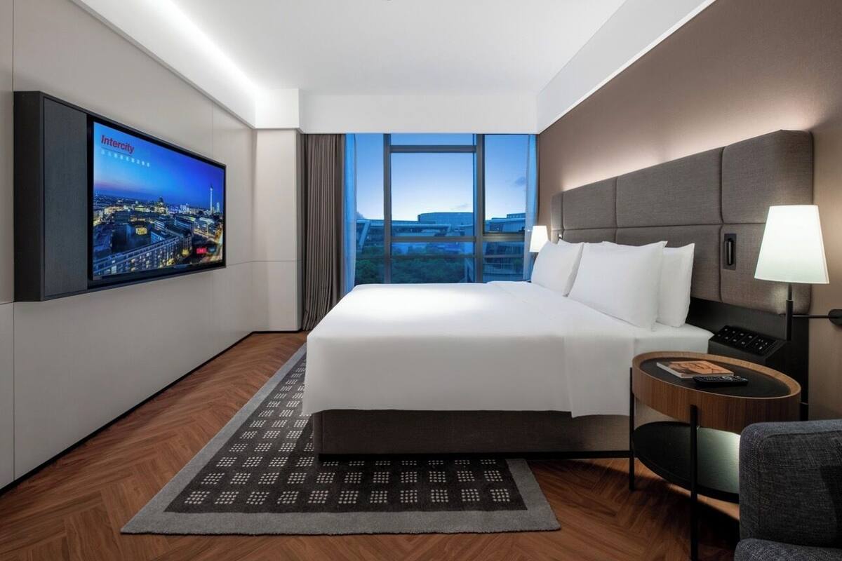 Une chambre d’hôtel moderne avec un grand lit, un téléviseur fixé au mur, une table de chevet et une vue sur la ville.