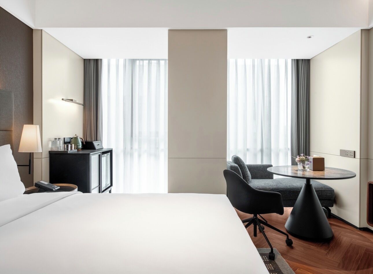 Une chambre d’hôtel avec un grand lit, un bureau avec une lampe, une chaise, une petite table avec un vase et une fenêtre avec des rideaux.