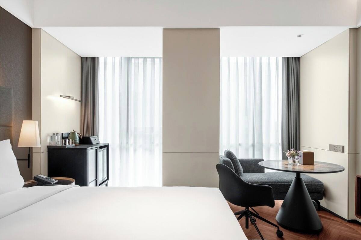 Une chambre d’hôtel avec un grand lit, un bureau avec une lampe, une chaise, une petite table avec un vase et une fenêtre avec des rideaux.