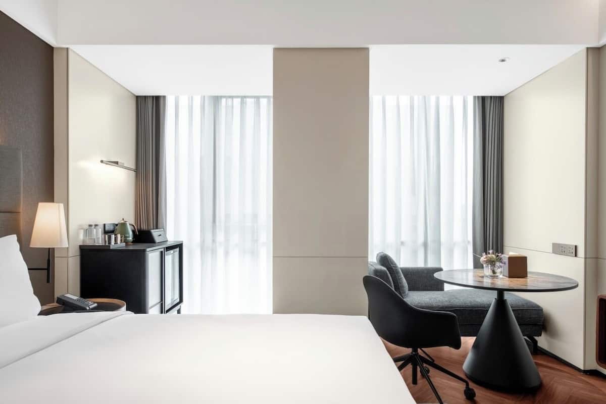 A hotel room with a large bed, a desk with a lamp, a chair, a small table with a vase, and a window with curtains.