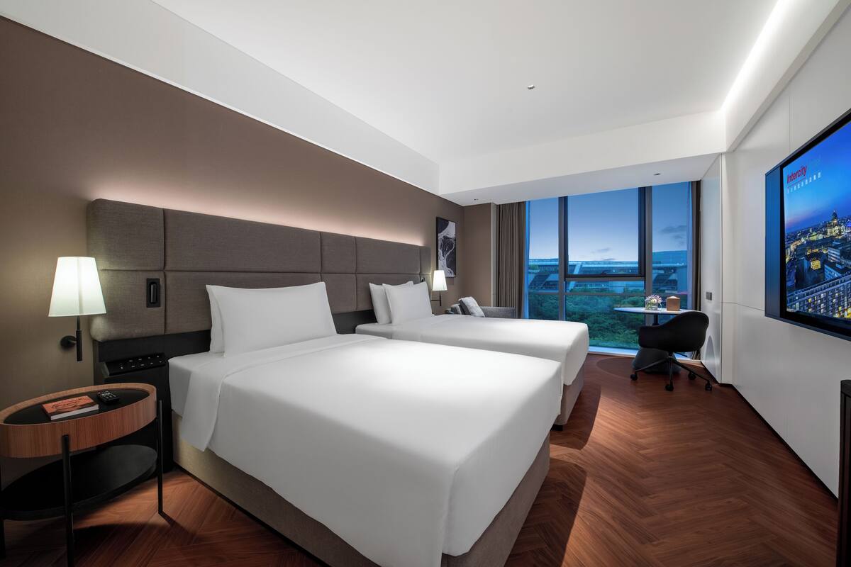 Une chambre d’hôtel avec deux lits, un bureau, une chaise, une télévision et une grande fenêtre donnant sur la ville.